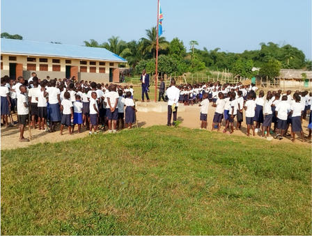 Students in uniforms gather for an assembly near a flagpole in a schoolyard.