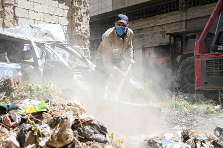 A person in protective gear shovels dirt and debris in a cluttered urban area.