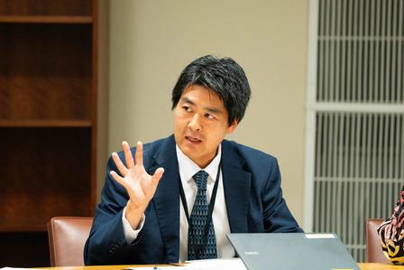 A man in a suit gestures while discussing at a conference table.