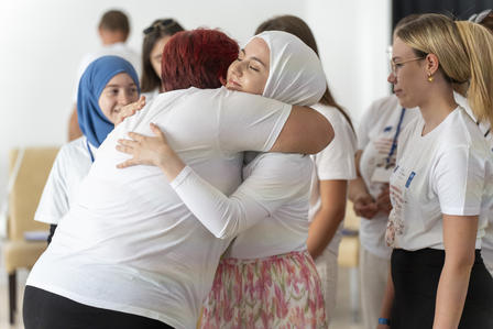 Two women embrace warmly, surrounded by others in casual attire, in a bright setting.