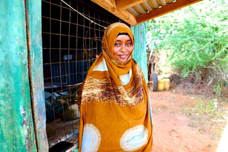 Woman in a brown patterned shawl standing outside a rustic building amidst greenery.