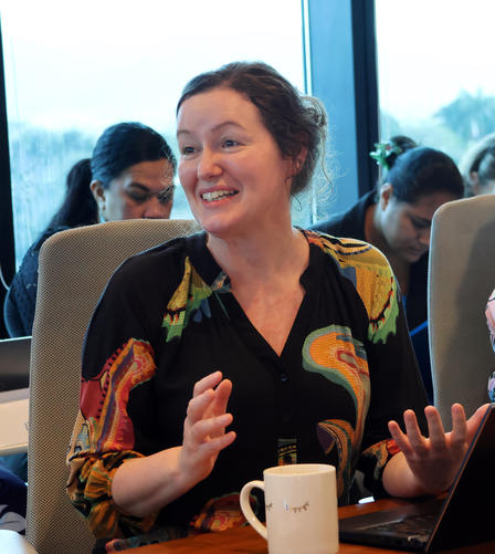 A smiling woman in a colorful blouse gestures while speaking at a meeting.