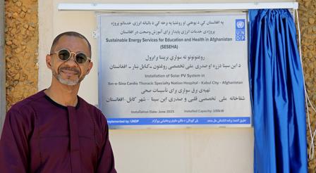 Man in traditional attire stands beside a plaque mounted on a wall.