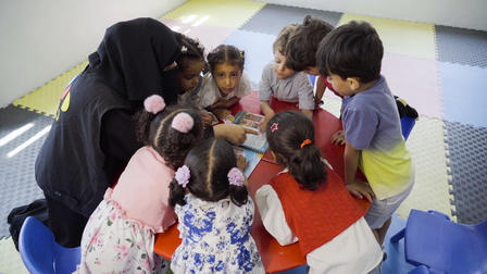 A teacher and several young children gather around a table, looking at a device together.