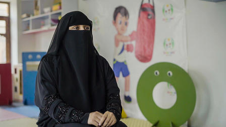 Woman in a black niqab seated in a play area, colorful background with a children's mural.