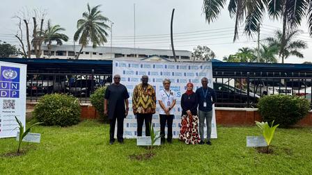 Five individuals stand in a grassy area with palm trees and a backdrop of a banner.