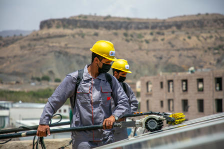 Two construction workers in yellow helmets and masks on a rooftop with mountains in the background.