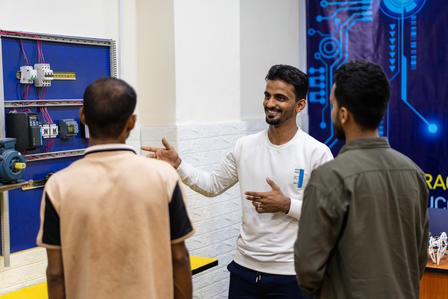 Three men engaged in discussion in a workshop, with equipment displayed on the wall behind them.
