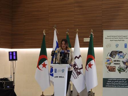 A woman speaks at a podium surrounded by flags and a banner in a modern conference room.
