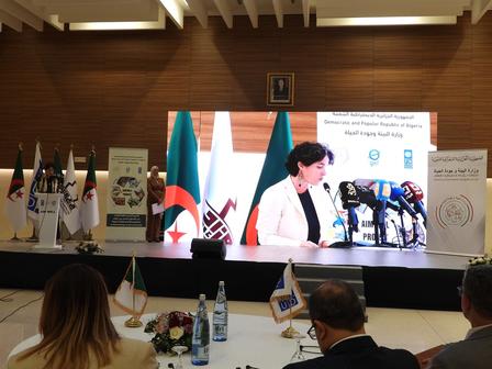 A woman speaks at a podium with several flags and a large screen behind her in a conference setting.