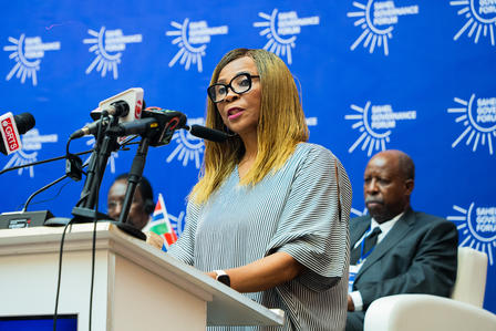 Woman speaking at a podium with microphones, a blue backdrop, and seated colleagues.