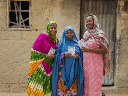 Three women dressed in colorful traditional clothing stand together, smiling and holding brochures.