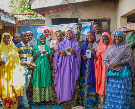 A group of diverse individuals stands together, smiling and holding cards in a colorful outdoor setting.
