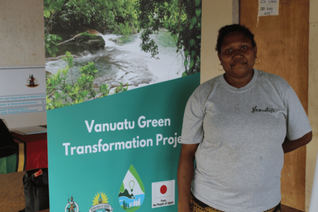 A woman stands beside a banner featuring a river and greenery for the Vanuatu Green Transformation Project.