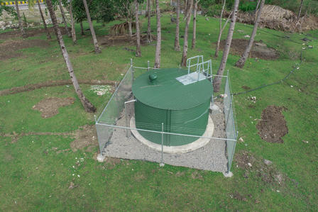 Green storage tank enclosed by a fence, situated on grassy land with trees.
