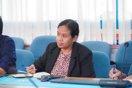 A focused woman in a black blazer sits at a table with a notebook and pen.