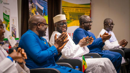 A group of four men in traditional and formal attire, sitting and clapping, in a conference setting.