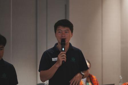 A man speaking into a microphone in a conference room, wearing a dark polo shirt.