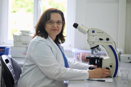 A woman in a lab coat sits at a desk with a microscope, smiling confidently.