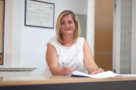 Smiling woman in a white blouse writes in a notebook at a desk.