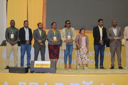 A diverse group of nine people standing on a stage, all wearing name tags and smiling.