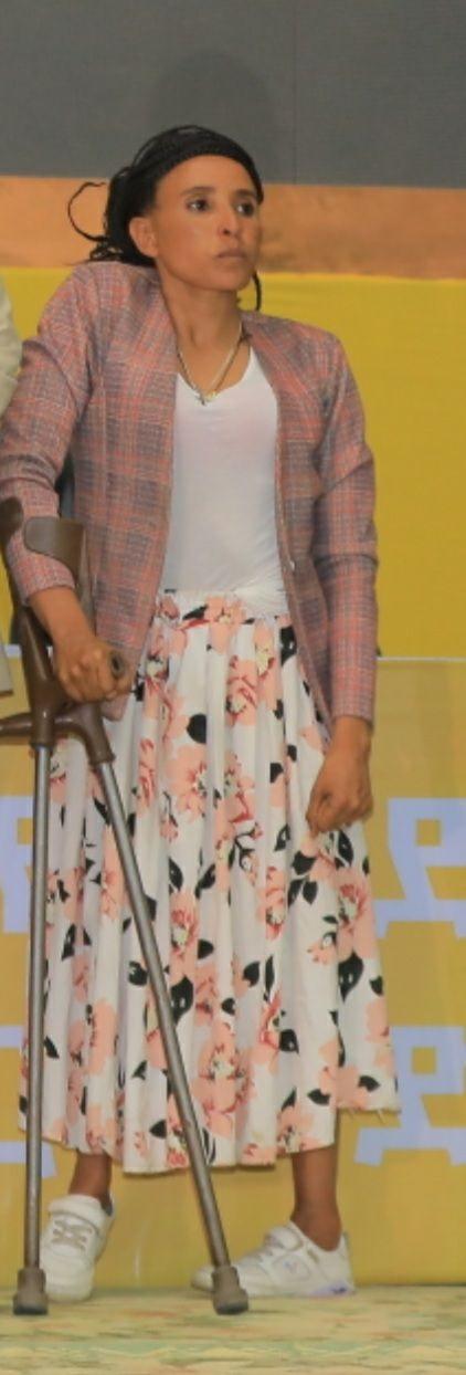 Woman in a floral skirt and plaid blazer stands with a cane on stage.