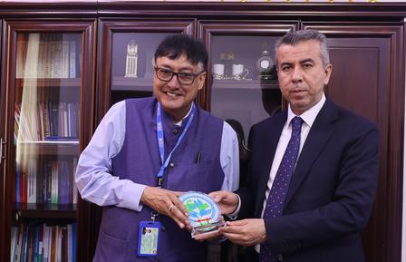 Two men pose for a photo, exchanging a decorative plate in an office setting.
