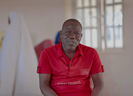 A man in a red shirt sits in a bright room, looking directly at the camera.