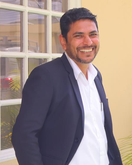 Smiling man in a suit stands near a window, with a light background and greenery.