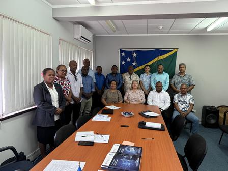 Group photo of diverse individuals standing in a conference room with a flag in the background.