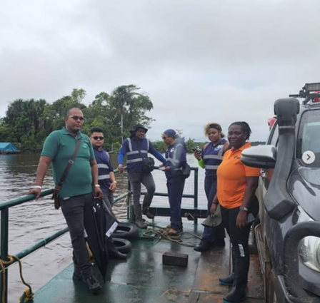 A group of seven people on a boat, surrounded by water and greenery, dressed in work attire.