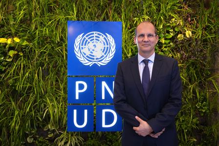 Man in a suit stands in front of a green wall, with a UN logo and PN/UD signage behind him.