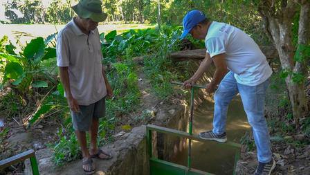 Two men by a water sluice; one adjusts the gate while the other observes in a lush green setting.