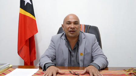 A man in a gray suit sits at a desk with a flag in the background.