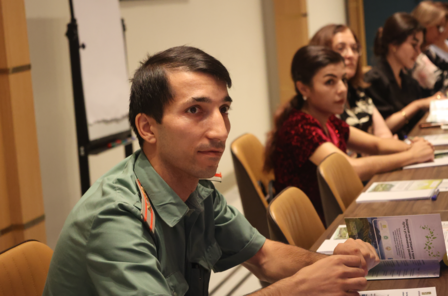 A young man in a green uniform sits at a conference table, focusing on a discussion.
