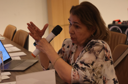 A woman speaks into a microphone during a meeting, gesturing with her hand.