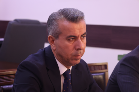 A man with short gray hair and a serious expression sits at a conference table.