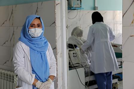 A healthcare worker in a blue hijab and mask stands confidently, while another tends to a patient.