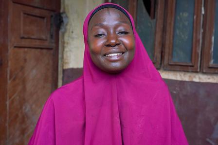 Smiling woman in a pink hijab standing outdoors near a rustic building.