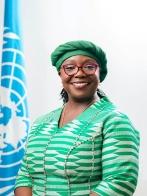 Smiling woman in green attire with a matching headwrap, standing by a UN flag.