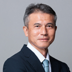 A man in a dark suit and light blue tie, smiling confidently against a gray backdrop.