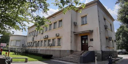 A large, light-colored building with multiple air conditioning units and large windows, surrounded by trees.