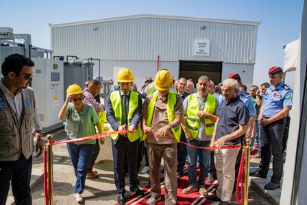 A group of people in safety vests and hard hats cuts a ribbon at a formal event outdoors.