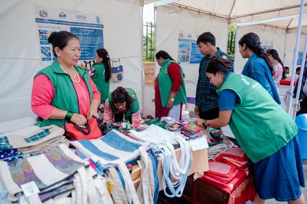 A group of people wearing green vests interact around a table filled with colorful handmade goods.