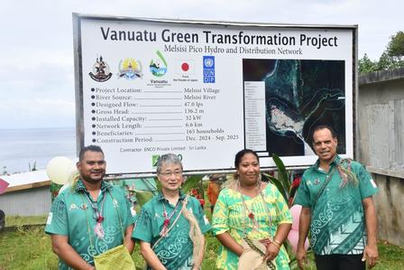 Four individuals in green attire stand in front of a project sign in a coastal setting.