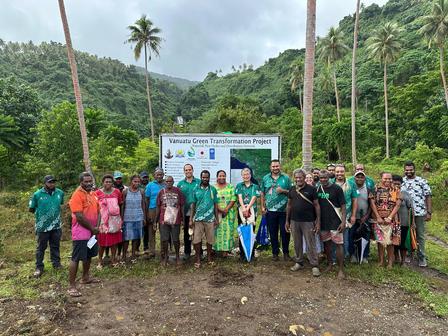 Group of people standing together outdoors near a sign, surrounded by palm trees and lush greenery.