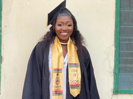 A smiling graduate in a black gown and cap, wearing a gold graduation sash, stands against a wall.