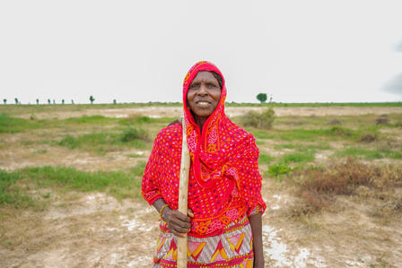 Woman in a red shawl smiles while holding a stick, standing in an open field.