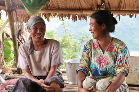 Two women sit under a thatched shelter by a lakeside; one holds yarn.