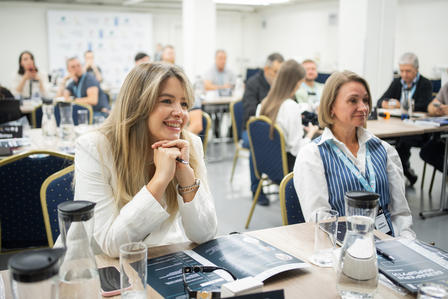 Two attendees at a conference table with their hands near their faces; others sit at surrounding tables.